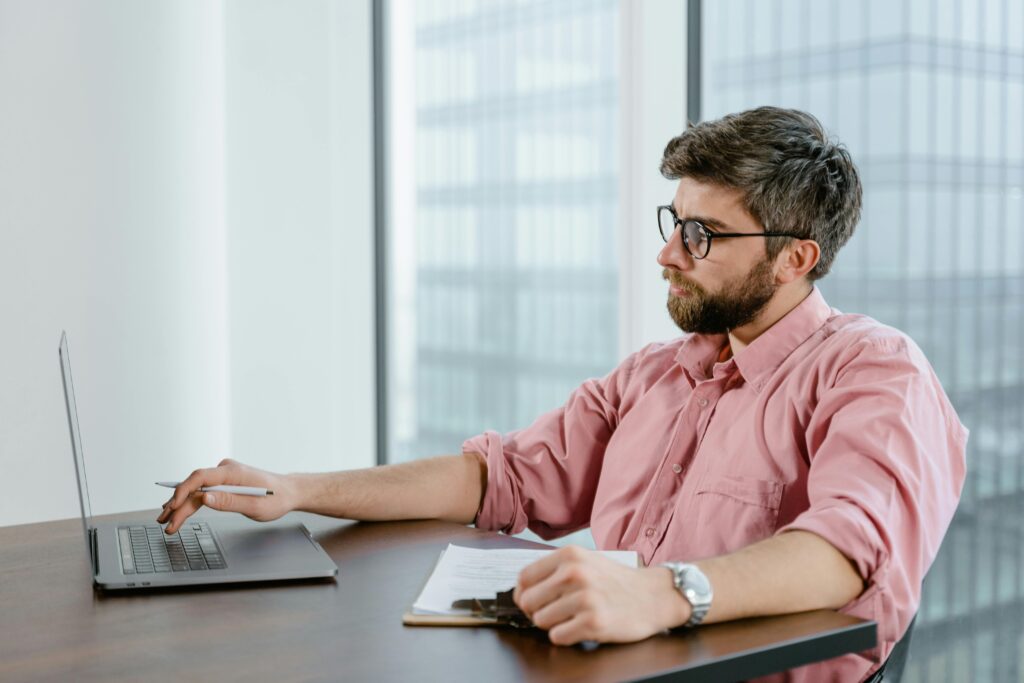 A focused businessman in a pink shirt uses a laptop at his office desk, with cityscape views.