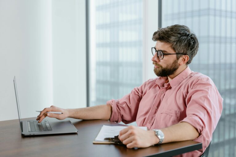 A focused businessman in a pink shirt uses a laptop at his office desk, with cityscape views.